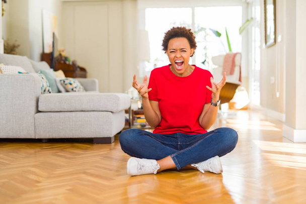 Nuori kaunis afrikkalainen amerikkalainen nainen istuu lattialla kotona hullu ja hullu huutaa ja huutaa aggressiivinen ilme ja kädet ylhäällä. Turhautumisen käsite
. - Valokuva, kuva