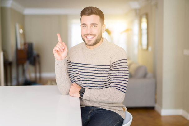Jóképű fiatalember visel egy pulóvert otthon, egy nagy mosollyal az arcán, a kéz és az ujjak mutasson a kamera oldalon. - Fotó, kép