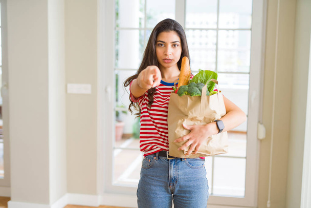 Mladá žena držící papírový sáček plný potravin ukazuje prstem do kamery a vám, rukou znamení, pozitivní a sebevědomý gesto z přední - Fotografie, Obrázek