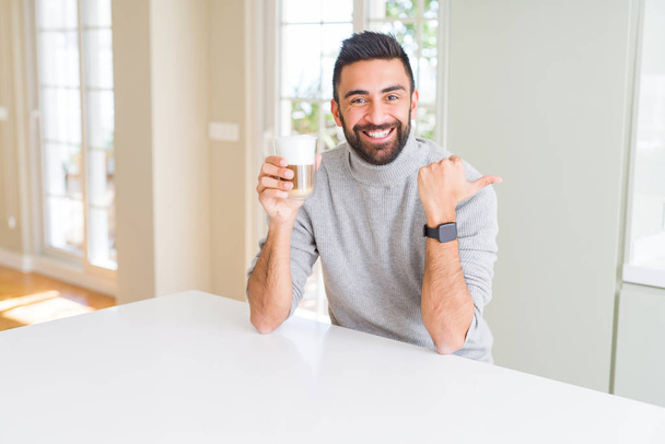 Przystojny mężczyzna hiszpanin picia filiżankę kawy, wskazując i Wyświetlono z kciukiem do boku z szczęśliwy twarz uśmiechający się - Zdjęcie, obraz