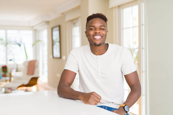 Όμορφος αφρικανική αμερικανική άνδρα στο λευκό τραπέζι στο σπίτι με μια ευτυχισμένη και δροσερό χαμόγελο στο πρόσωπο. Τυχερό πρόσωπο. - Φωτογραφία, εικόνα