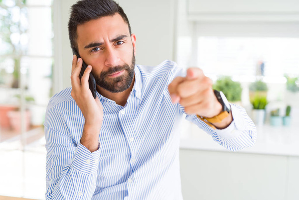 Handsome hispanic business man having a conversation talking on smartphone pointing with finger to the camera and to you, hand sign, positive and confident gesture from the front - Photo, Image