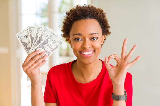 Joven mujer afroamericana sosteniendo billetes de banco de veinte dólares haciendo ok signo con los dedos, excelente símbolo
 - Foto, Imagen