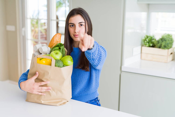Hermosa mujer joven sosteniendo bolsa de papel llena de alimentos saludables señalando con el dedo a la cámara y a usted, signo de mano, gesto positivo y seguro de la parte delantera
 - Foto, imagen