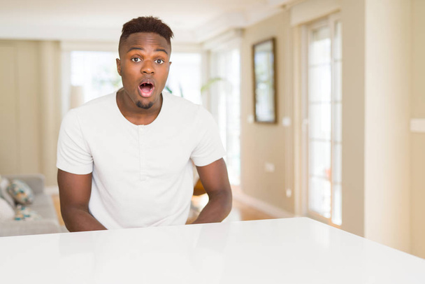 Bel homme afro-américain sur la table blanche à la maison effrayé et choqué par l'expression surprise, la peur et le visage excité
. - Photo, image
