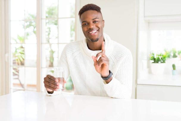Hombre afroamericano goteando un vaso de agua fresca sorprendido con una idea o pregunta apuntando con el dedo con la cara feliz, número uno
 - Foto, Imagen
