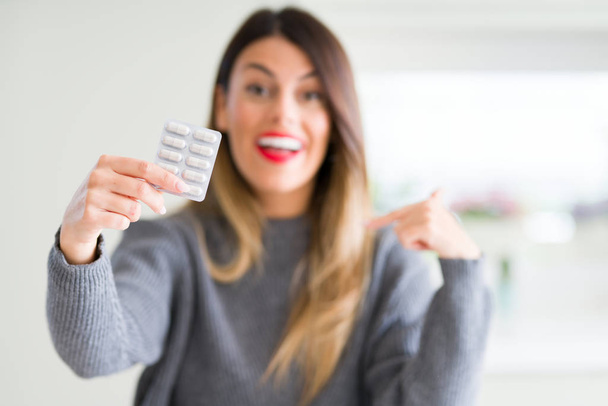 Молодая красивая женщина держит дома фармацевтические таблетки с неожиданным лицом, указывающим пальцем на себя
 - Фото, изображение