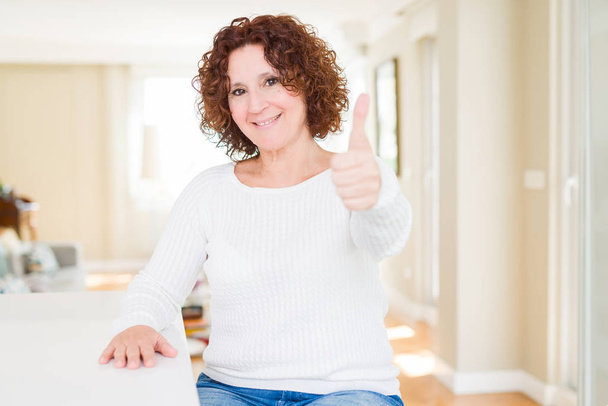 Krásná starší žena nosí bílý svetr doma dělá šťastné palec nahoru gesto rukou. Schvalování výraz při pohledu na fotoaparát ukazuje úspěch. - Fotografie, Obrázek