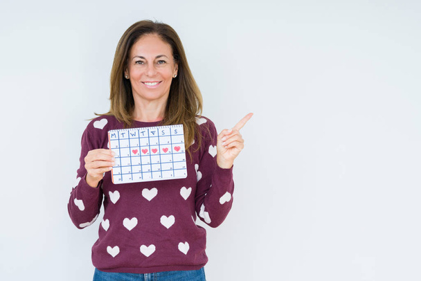 Жінка середнього віку, що тримає календар менструацій над ізольованим тлом, дуже щаслива, вказуючи рукою і пальцем на бік
 - Фото, зображення