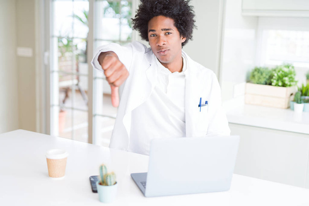 Médico afro-americano trabalhando com laptop na clínica com rosto irritado, sinal negativo mostrando antipatia com os polegares para baixo, conceito de rejeição
 - Foto, Imagem