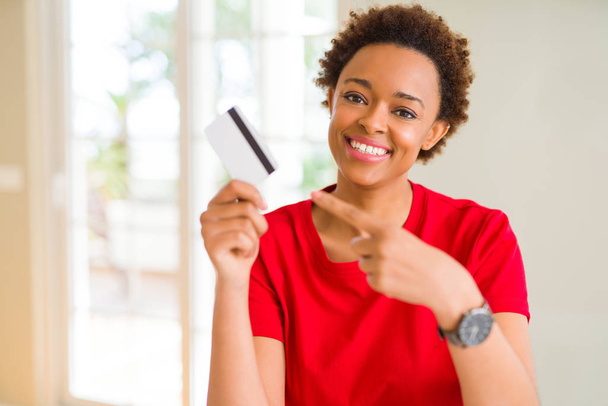 Jovem afro-americana segurando cartão de crédito muito feliz apontando com mão e dedo
 - Foto, Imagem