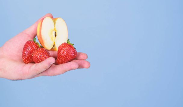 Help yourself. Fresh harvest of ripe fruit berries. Hand proposes take strawberry and apple fruit. Male hand with strawberries and half of apple blue background. Fruits organic treat or snack - Photo, image