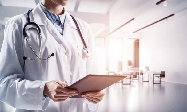 Close up image of young female medical employee in white suit using tablet while standing inside white hospital building. Medical industry concept - Foto, Imagem