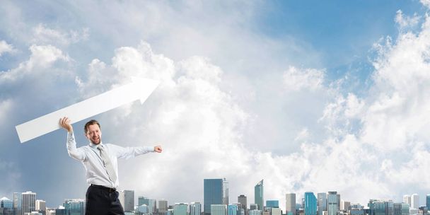 Poziome ujęcie młodego biznesmena rzucającego wielką białą strzałą w powietrze stojącego naprzeciw nowoczesnego widoku miejskiego i zachmurzonego nieba na tle - Zdjęcie, obraz