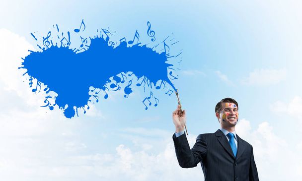 Foto horizontal de joven y alegre hombre de negocios en traje negro de pie con salpicadura musical de color azul contra el paisaje nublado en el fondo
. - Foto, Imagen