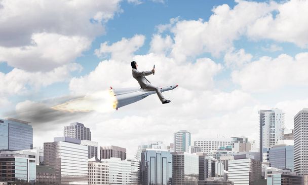 Image conceptuelle de jeune homme d'affaires en costume volant sur fusée avec paysage urbain moderne avec gratte-ciel et ciel bleu en arrière-plan
. - Photo, image