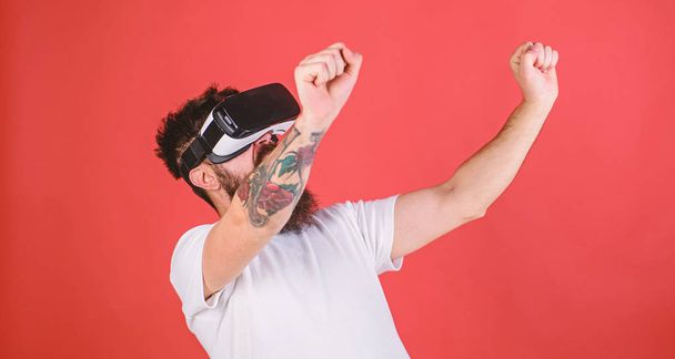 Man with beard in VR glasses driving motorbike, red background. Virtual driving lessons concept. Hipster on excited face driving bike on high speed in virtual reality with modern digital gadget - Photo, image