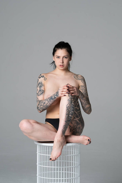 fiatal gyönyörű lány tetoválás stúdió ül egy széken, fekete fehérnemű meztelenül pózol - Fotó, kép