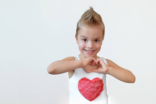 Lovely child boy smiling shows heart sign. Portrait kid blond 6 year with smile making hands gesture love. Studio isolated white background. - Foto, Imagem