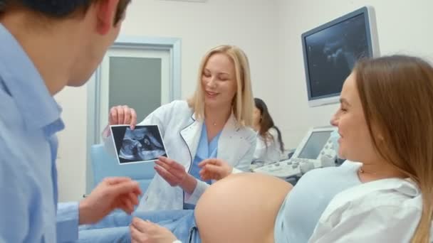 Médico femenino mostrando la ecografía de la mujer embarazada en la foto explicándolo y sonriendo
 - Imágenes, Vídeo