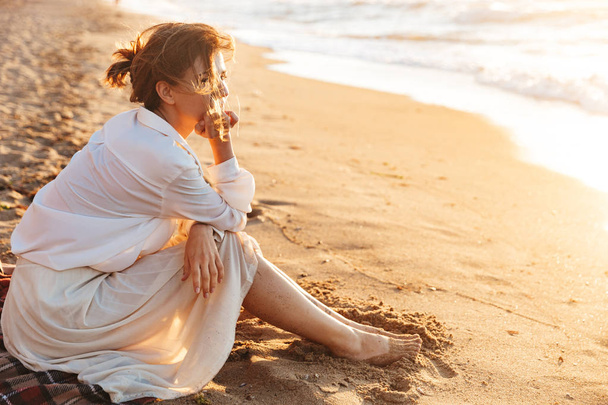Image of joyful woman 20s sitting on sand and looking at sea, wh - Valokuva, kuva