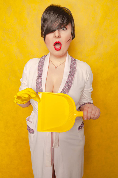 plump woman with short black hair stands in a pink Bathrobe with a rubber cleaning glove on her hand and a dustpan on a yellow Studio background - Photo, Image