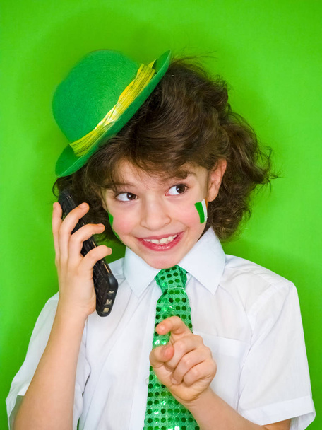 Funny St Patricks day little boy wearing a green hat and shamrock, the Irish flag on his cheeks talking on the phone.A child listening to good news on the phone is incredibly excited and joyful - Foto, Imagem