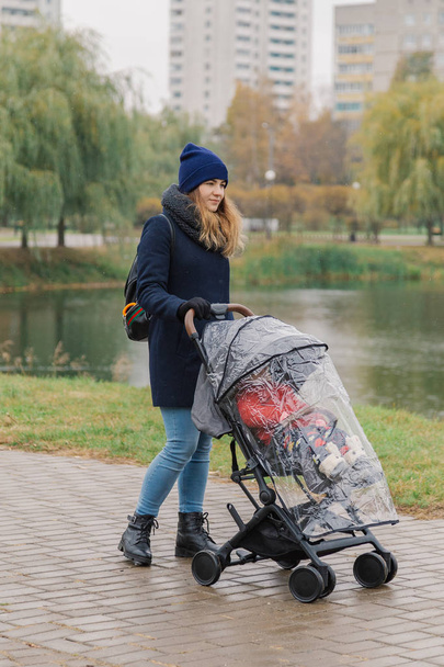 Una mujer camina en el parque con un cochecito y un niño pequeño cerca del lago
. - Foto, Imagen
