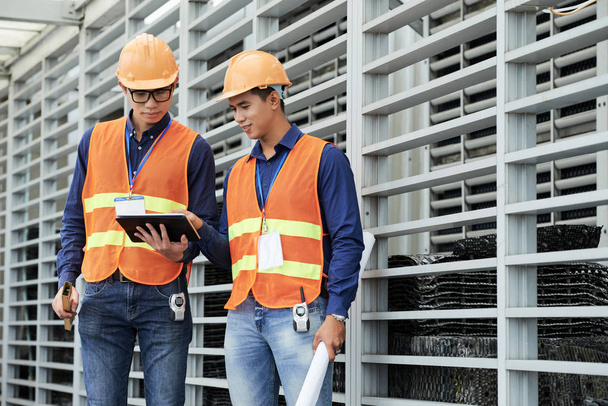 Dois homens asiáticos jovens em hardhats e coletes de navegação tablet enquanto de pé perto da parede do edifício em construção
 - Foto, Imagem
