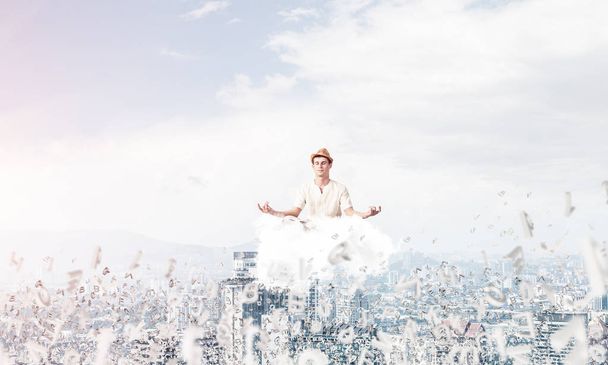 Man in white clothing keeping eyes closed and looking concentrated while meditating on cloud among flying letters with cityscape view on background. - Фото, изображение