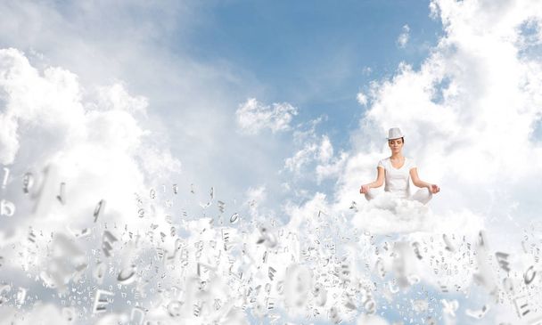 Woman in white clothing keeping eyes closed and looking concentrated while meditating among flying letters in the air with cloudy skyscape on background. - Photo, image