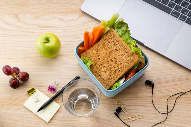 healthy sandwich in plastic container with fruits and office items next to opened laptop on wooden table. Takeaway food for lunch at work  - Photo, image