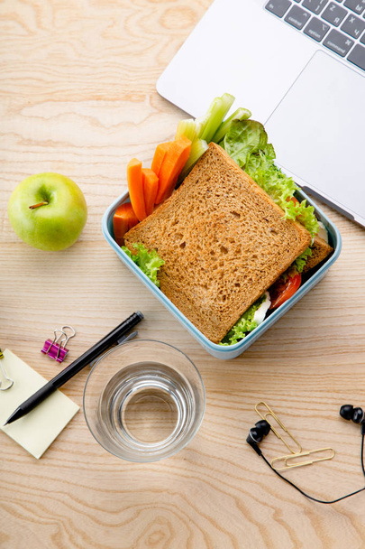 healthy sandwich in plastic container with fruits and office items next to opened laptop on wooden table. Takeaway food for lunch at work  - Foto, Imagem