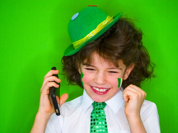 Funny St Patricks day stunning little boy wearing a green hat and shamrock, the Irish flag on his cheeks talking on the phone. - Foto, immagini