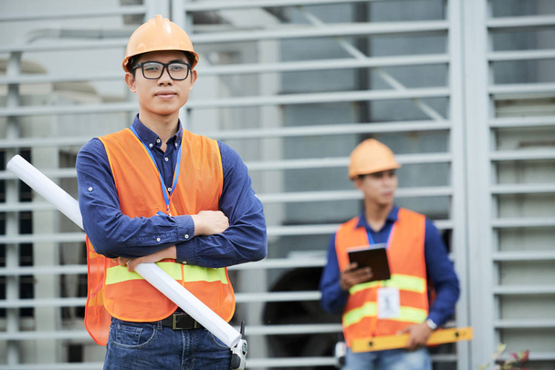 Aantrekkelijke Aziatische man in de helm en gilet ontwerp houden en kijken naar de camera terwijl staande op de bouwplaats in de buurt van collega - Foto, afbeelding