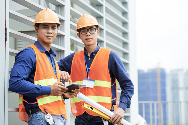 Deux jeunes hommes asiatiques penchés sur le mur de construction et regardant la caméra tout en se tenant debout sur le chantier de construction
 - Photo, image