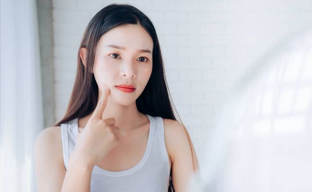 Young Asian woman squeeze acne problem face looking at mirror her without Skincare.  - Foto, afbeelding