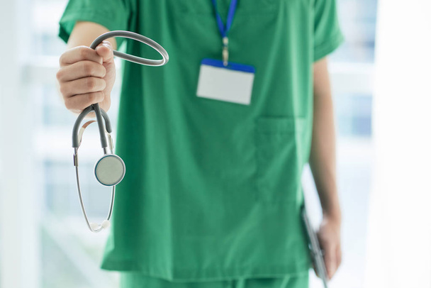 Gewas man in groene medische uniforme weergegeven: stethoscoop naar camera terwijl staande op onscherpe achtergrond van ziekenhuis hall - Foto, afbeelding