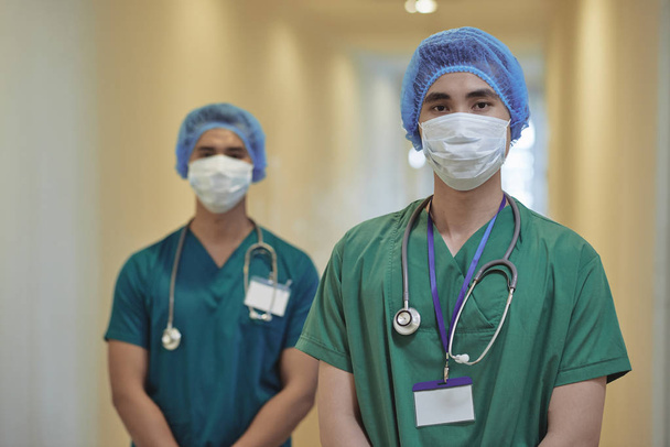 Δύο Ασίας άνδρες σε χειρουργό ομοιόμορφη βλέπουν φωτογραφική μηχανή, ενώ στέκεται στην θολή φόντο από διάδρομο νοσοκομείο - Φωτογραφία, εικόνα