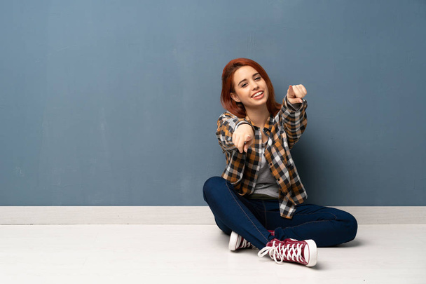 Nuori punapää nainen istuu lattialla osoittaa sormella sinua hymyillen
 - Valokuva, kuva