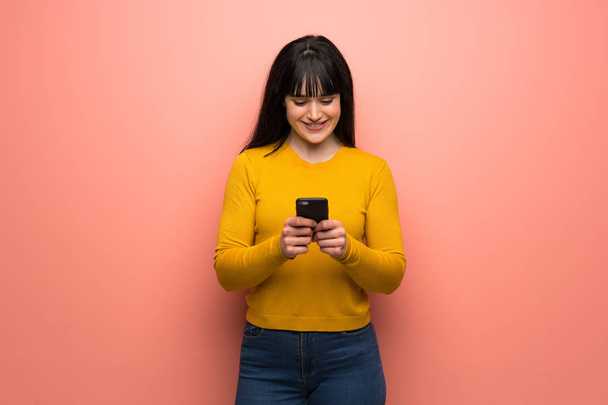 Жінка з жовтим светром над рожевою стіною надсилає повідомлення з мобільним
 - Фото, зображення