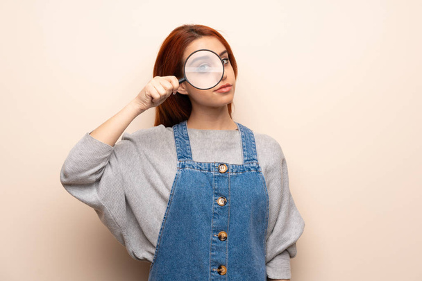 Young redhead woman over isolated background taking a magnifying glass and looking through it - Photo, image