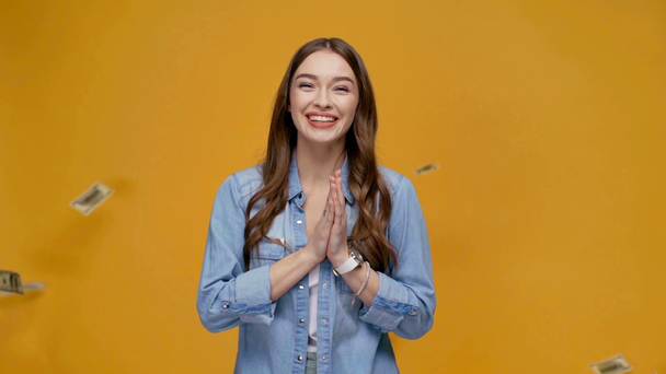 beautiful happy girl doing please gesture and looking at falling dollar banknotes in slow motion isolated on yellow - Footage, Video