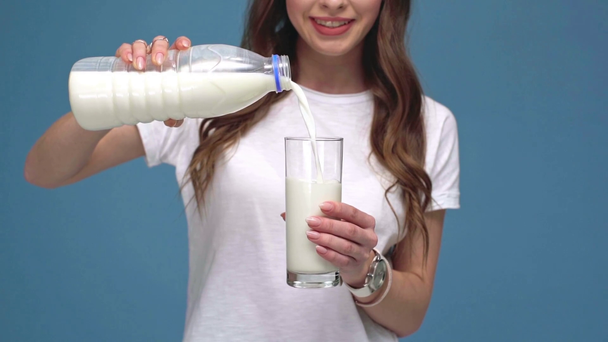 Kız dökülen süt yavaş çekimde, gülümseyerek ve mavi izole kamera cam gösterilen kırpılmış görünümü - Video, Çekim
