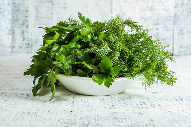 Green parsley and dill in a white plate on a white wooden table - Foto, Bild