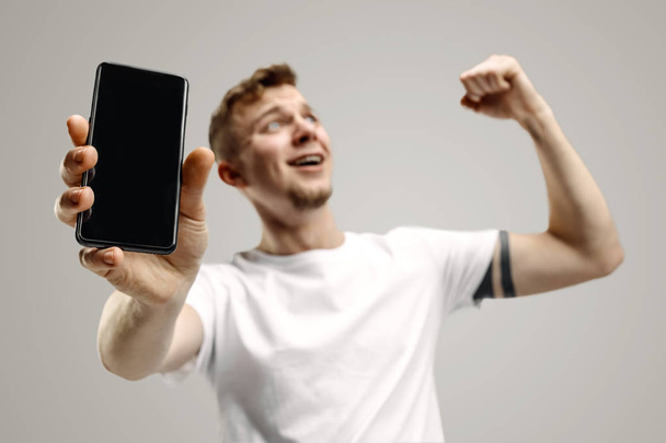 Young handsome man showing smartphone screen isolated on gray background in shock with a surprise face - Foto, immagini