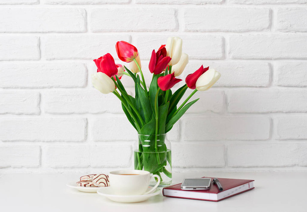 tulip flowers are in a vase on the table, cup of coffee, diary and smartphone, white brick wall as background - Zdjęcie, obraz