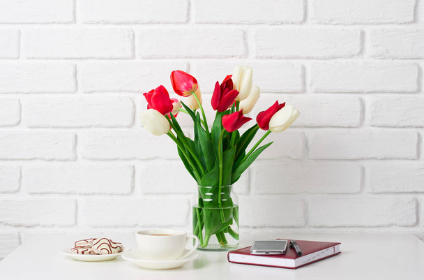 tulip flowers are in a vase on the table, cup of coffee, diary and smartphone, white brick wall as background - Photo, Image