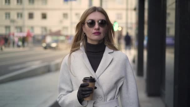 Attractive young woman in white coat drinking coffee walking - Πλάνα, βίντεο