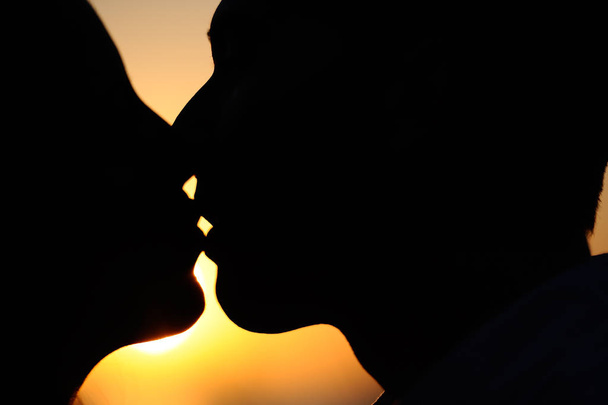 silhueta de faces laterais beijando em um fundo por do sol de cores alaranjadas
 - Foto, Imagem